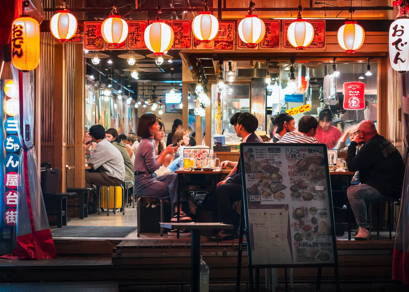 In a cozy Japanese Izakaya, people exchange greetings before saying itadakimasu and enjoying their meal, seated around a wooden table.